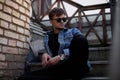 Cool young man hipster in stylish blue denim clothes in trendy sunglasses sits on a vintage staircase near brick wall in the city Royalty Free Stock Photo