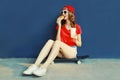 Cool young woman with burger and coffee cup sitting on skateboard wearing baseball cap, sunglasses over blue wall Royalty Free Stock Photo