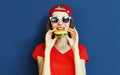 Cool young woman eating burger wearing baseball cap, sunglasses over blue wall Royalty Free Stock Photo