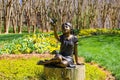 A cooper statue of a little girl holding a butterfly surrounded by lush green grass, bare winter trees, colorful flowers