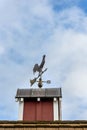 Copper rooster weathervane on top of red rooftop cupola with a blue sky and white clouds in the background