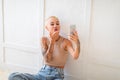 Coquette lady with short haircut taking selfie, blowing air kiss to smartphone camera, sitting over white wall Royalty Free Stock Photo