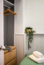 A corner of bedroom with part of bed with towels, green plant and openstyle wardrobe with hangers.