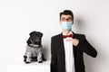 Coronavirus, pets and celebration concept. Amazed young man in face mask and suit pointing at cute black dog sitting