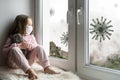 Coronavirus and quarantine concept, sad kid in medical mask looks out window, little girl sits on windowsill safe from corona