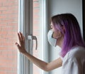 Coronavirus. Young girl with purple hair looking through the window and wearing mask protection. Quarantine.