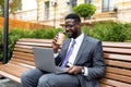 Corporate african american executive in formal wear sitting on bench near office center with laptop and takeout coffee