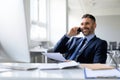 Corporate communication. Happy middle aged male manager talking on mobile phone, sitting at desk in front of computer Royalty Free Stock Photo