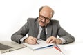 Corporate portrait bald 60s happy and friendly business man working writing on notepad at computer laptop office desk