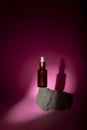A cosmetic bottle with a pipette balances on a gray stone against a pink background. Directional light falls on the jar