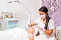 A cosmetologist in a medical mask and gloves holds a bowl of cosmetics, applying a brush mask on the client& x27;s face.