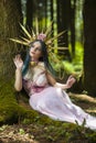 Costume Play. Magnificent Crowned Forest Nymph with Flowery Golden Crown Posing in Summer Empty Forest Near Tree Stem