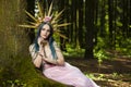 Costume Play. Magnificent Crowned Forest Nymph with Flowery Golden Crown Posing in Summer Empty Forest Near Tree Stem