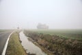 Country road bordered by fields and a channel with an abandoned house in the distance on a foggy day in winter Royalty Free Stock Photo
