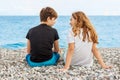 Couple of beautiful teens, first love. Guy hugs a girl sitting on the pebble beach next to each other and looking at the sea.