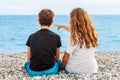 Couple of beautiful teens, first love. Guy hugs a girl sitting on the pebble beach next to each other and looking at the sea.