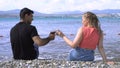 Couple drinking wine on stone beach. Media. Young man and woman drink wine sitting on rocky shore near blue sea. Young