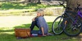 Adult woman sitting on plaid blanket by pond in park with picnic basket and bicycles Royalty Free Stock Photo