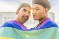 Couple of gay men in love embracing each other, wrapped in a gay flag, in a park at sunset, with reflections of the sun. Royalty Free Stock Photo