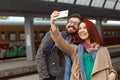 Couple of hipster travelers photographing a selfie with a smartphone in a train station. Travel concept. Mobile