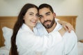 Couple Hugging Wearing White Bathrobes Posing Sitting In Bedroom Indoors