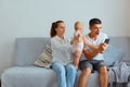 Couple with infant kid sitting on sofa in living room, mother giving baby to father, trying to attract his attention, amazed man Royalty Free Stock Photo