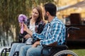 A couple of invalids on wheelchairs met in the park. A man is giving a bouquet of flowers to a woman.