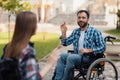 A couple of invalids on wheelchairs met in the park. A man is greeting a woman.