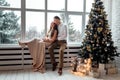 Couple in love in festive clothers sitting and hugging near the large window and Christmas tree in decorated studio. happy holiday