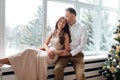 Couple in love in festive clothers sitting and hugging near the large window and Christmas tree in decorated studio. happy holiday