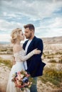 Couple in love hugs and kisses in fabulous mountains in nature. Girl in long white dress with bouquet of flowers in her hands, man Royalty Free Stock Photo