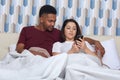 Couple in love lieing on white bed. Young darkskinned man and woman looking at mobile phone, reading or watching something