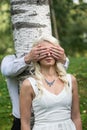 Couple in love. Man covered eyes of smiling blonde woman by his hands in park.