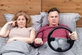 The couple is lying in bed, the man is holding the steering wheel and shifting gears, the woman is in shock and twists her finger