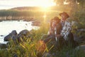 Couple man and woman in felt hats wrapped in whool blanket sitting near lake on sunset. Weekend picnic on nature, local travel,