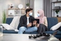 Couple in masks sitting on sofa at home. Young Arabian Muslim couple, spending time during desease and isolation at home