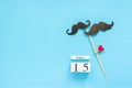 Couple paper mustache props fastened clothespin heart and calendar 15 August on blue background. Concept Homosexuality gay love.