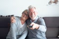 Couple of pensioners seniors looking and watching the TV together at home sitting on the sofa - two mature people smiling and man Royalty Free Stock Photo