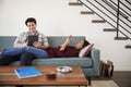 Couple Relaxing On Sofa At Home Using Mobile Phone And Digital Tablet