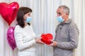 Couple seniors celebrate Valentine& x27;s Day in mask. Man gives a woman gift box in shape of a heart. Romantic relationships