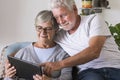 Couple of seniors smiling and looking at the same tablet hugged on the sofa - indoor, at home concept - caucasians mature and Royalty Free Stock Photo