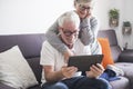 Couple of seniors smiling and looking at the same tablet hugged on the sofa - indoor, at home concept - caucasians mature and Royalty Free Stock Photo