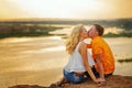 Couple sitting on bank of river at sunset. Kiss.