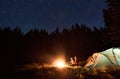 Couple sitting by burning fire near tent under bright starry sky in pine forest. Romantic night camping.