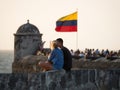 Couple sitting on city wall with colombian national flag waving in wind on Bastion of Santo Domingo Cartagena Colombia