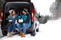 Couple sitting in open car trunk full of luggage near road, space for text. Winter