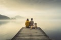 Couple sitting on a wooden jetty on a lake in the morning, A family with a small yellow dog resting on a pier and looking at the Royalty Free Stock Photo
