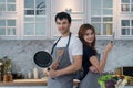 Couple standing back to back in kitchen. Smiling handsome young man wear an apron while holding frying pan and wooden spoon Royalty Free Stock Photo