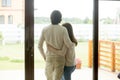 Couple standing at home, embracing looking outside, rear view