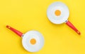 Couple of symmetric white ceramic frying pans with red handle on bright yellow background. Sunny side up fried eggs